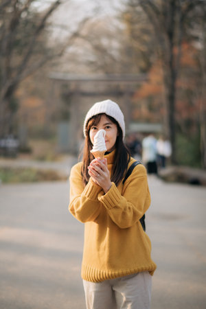 A young woman in yellow sweater enjoying ice cream outdoors, cheerful and relaxed, autumn park background, asian ethnicity, holiday vibeの写真素材