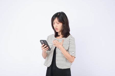 A southeast asian woman in striped jacket holding smartphone with worried expression standing against white background, emotional reaction to message or newsの写真素材
