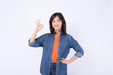 A southeast asian woman in casual denim shirt and orange top smiling confidently while making okay hand gesture on white backgroundの写真素材