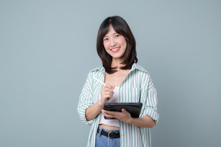 A young asian woman teacher or student holding digital tablet and pencil, smiling, taking notes, standing over green backgroundの写真素材