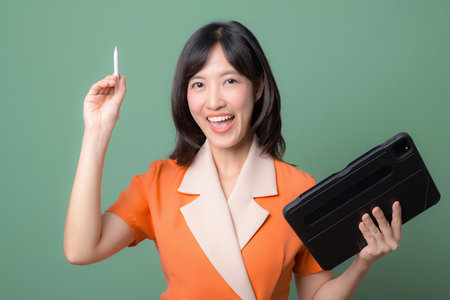 A southeast asian woman in orange suit holding tablet and stylus smiling confidently against green backgroundの写真素材