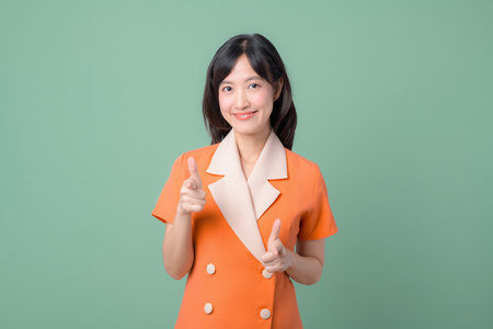A southeast asian woman in orange blazer smiling confidently and pointing fingers forward against green background, expressing cheerful and positive emotion in studio portraitの写真素材