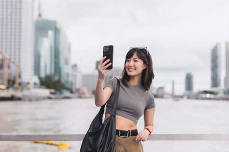 A solo southeast asian woman teenager blogger world travel. local cultural tourism experience concept for explorer bangkok thailand. express joy with smartphone, wait for friend boat chao phrayaの写真素材
