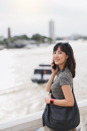 A happy solo southeast asian woman on world travel tourism in bangkok thailand. great local cultural experience for young explorer using smartphone near boat on chao phraya riverの写真素材