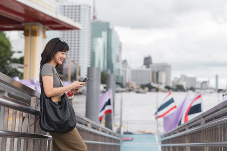 A happy young southeast asian woman searching map on smartphone tourism guide. world travel explorer enjoying local cultural experience in bangkok thailand by chao phraya riverの写真素材