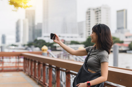 A happy solo southeast asian woman teenager blogger on world travel express joy take photo with smartphone by chao phraya river bangkok. local cultural tourism experience in asia for an explorerの写真素材