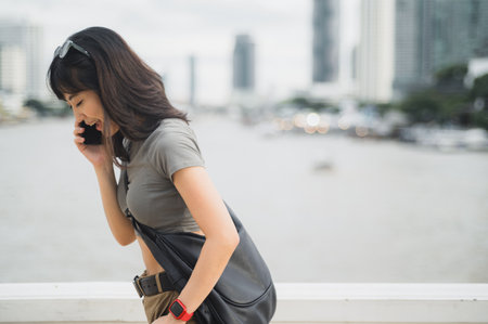 A solo southeast asian woman, teenager world travel explorer, on tourism trip in asia. blogger has serious phone call with friend via smartphone by chao phraya river in bangkok, thailandの写真素材
