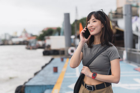 A happy solo southeast asian woman, teenager blogger, on world travel tourism experience in thailand. she wait for boat on chao phraya river in bangkok using smartphone to express joy to friendの写真素材