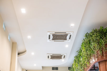 A modern white ceiling with cassette type air conditioner for cool comfort. indoor ventilation system provides clean air and climate control alongside recessed spotlightsの写真素材
