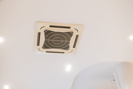 A cassette type air conditioner on white ceiling providing cool air for indoor climate control. ventilation system equipment offers comfort inside modern office interiorの写真素材