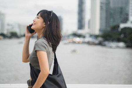 A happy solo southeast asian woman teenager travel blogger uses smartphone. an explorer enjoying local cultural experience asia waiting for friend or express boat by chao phraya river bangkokの写真素材