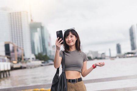 A happy solo southeast asian woman teenager blogger on world travel tourism trip in asia. explorer uses smartphone to express local cultural experience by chao phraya river in bangkok thailandの写真素材