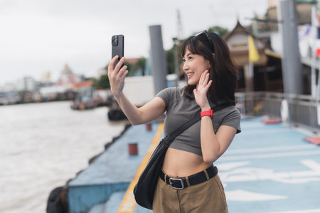 Happy solo southeast asian woman blogger express joy. Local cultural tourism concept experience for world travel explorer with smartphone wait for friend boat Chao Phraya river Bangkok Thailandの写真素材