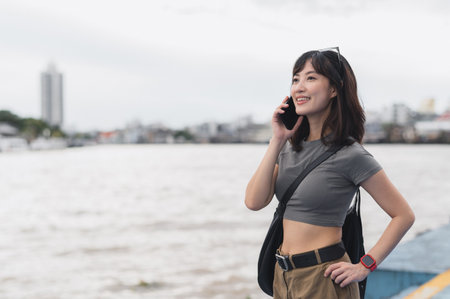 Happy solo Southeast Asian woman teenager blogger on world travel tourism waits for boat using smartphone to express joy to friend by Chao Phraya River in Bangkok Thailandの写真素材