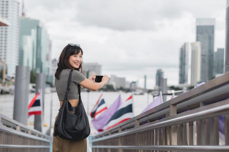 Happy solo Southeast Asian woman teenager blogger enjoy world travel tourism. Young explorer take photo with smartphone at Chao Phraya River Bangkok for local cultural experience in Asia Thailandの写真素材