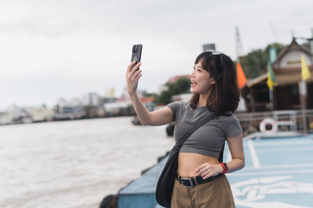 Happy solo Southeast Asian woman teenager blogger and world travel explorer on local cultural experience in Bangkok Thailand. Using smartphone on boat on Chao Phraya river. Asia tourism conceptの写真素材