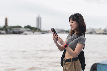 Happy solo southeast asian woman teenager blogger world travel tourism. Explorer wait for friend or express boat by Chao Phraya river. Local cultural experience smartphone concept Bangkok Thailandの写真素材