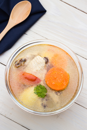 Bowl of chicken soup and chicken wings with carrots, potatoes and tomatoes on gray table mat with wooden spoonの写真素材