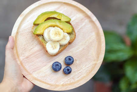 Hand holding wooden plate avocado  toast with banana honey and blueberry for healthy breakfast or snack.の写真素材