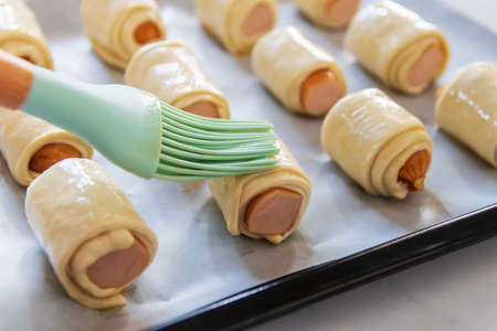Mini sausage wrapped in puff pastry in oven tray prepare for bake. Beating the egg nicely and brush the top with it to get the shiny and smooth top.の写真素材
