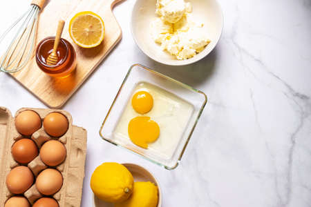 Baking ingredients on white table with copy space; top view. Eggs, flour, honey, lemon, cream, cream cheese and kitchen utensils on table.の写真素材