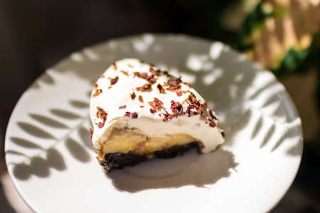 Banoffee pie with banana, whipped cream and caramel sauce on a white plate with sunlight and shadow from a tree in garden background.の写真素材