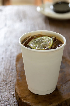 Paper cup of espresso mixed with lime juice and honey topped with sliced lime. Iced coffee cup on wooden table with soft sunlight.の写真素材