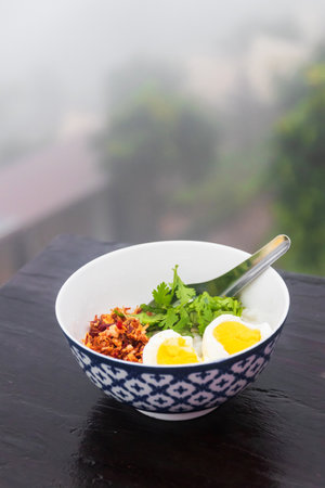 Bowl of rice congee and boiled eggs with fried garlic, chili and herbs. Breakfast with morning fog in rainy season.の写真素材