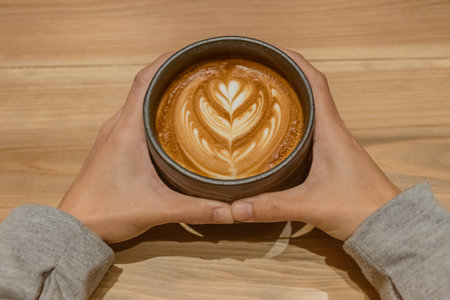 Cup of coffee with latte art on wooden table, women hold in hands.の写真素材