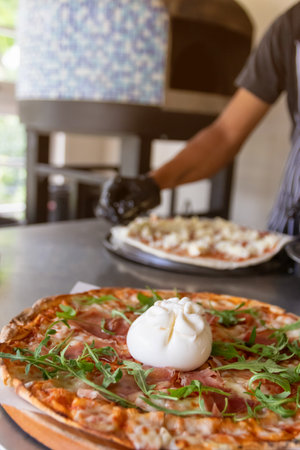 Burrata cheese topping on Italian pizza with prosciutto and arugula, served on the wooden board with traditional oven on background.の写真素材