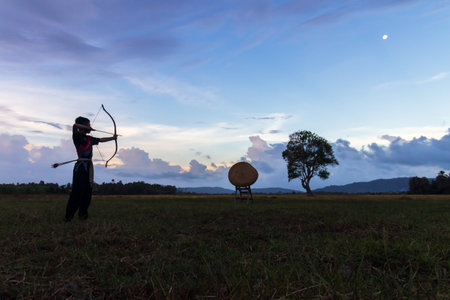 A man shooting an arrow at a target. The man is dressed in traditional clothing and the background...