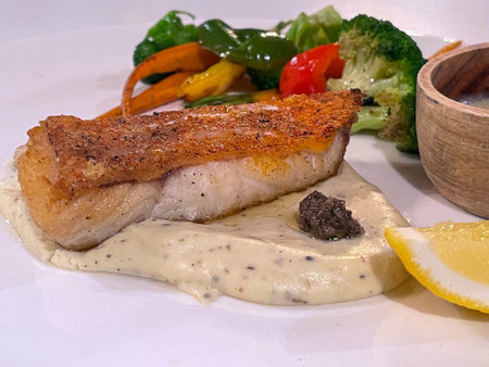 Delicious grilled fish fillet with creamy sauce, fresh vegetables, and a lemon slice, perfect for a...