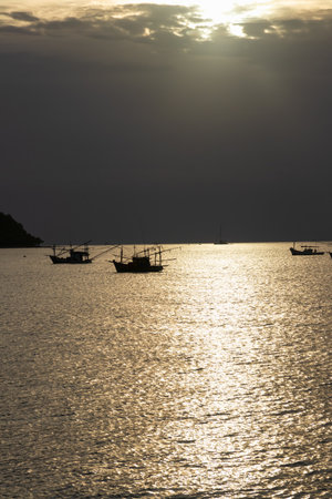 A serene sunset over calm waters, with fishing boats silhouetted against a dramatic sky, creating a tranquil coastal scene.の写真素材