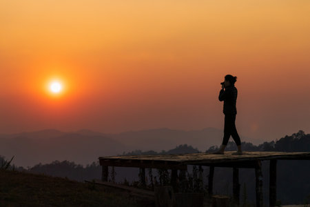 The photo shows a silhouette of a person standing and holding a camera in hands on a wooden walkway...