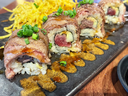 A seared beef sushi roll with rice, avocado and chive garnish on black plate, savory glaze and shredded garnish creating appetizing moodの写真素材