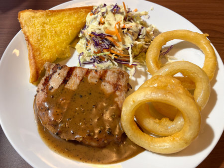 A grilled pork chop with pepper sauce, coleslaw, toast and golden onion ring offering comforting hearty mealの写真素材