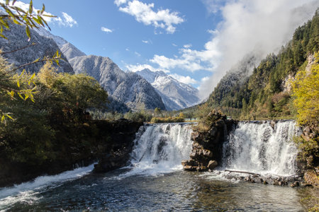 A snowy mountain waterfall in autumn valley with flowing river, evergreen forest and golden foliage at Bipenggou, China.の写真素材