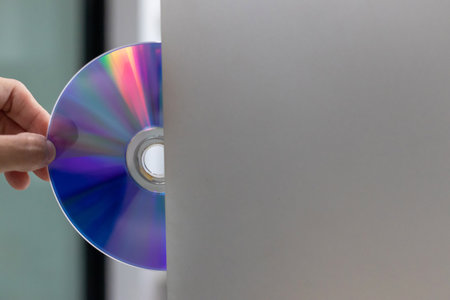 A hand holding optical disc next to silver laptop slot, colorful reflection and modern technology conceptの写真素材