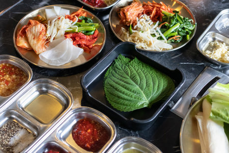 A green perilla leaf black tray with assorted korean banchan including kimchi, pickled radish, bean sprouts and dipping saucesの写真素材