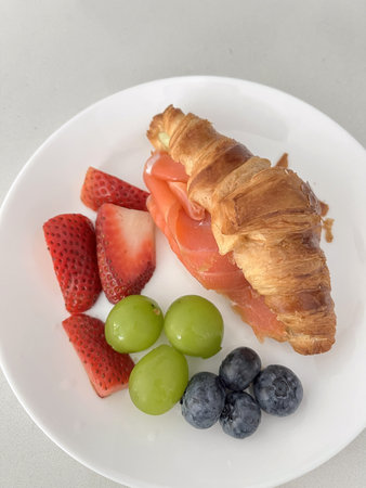 A smoked salmon croissant with fresh strawberries grapes and blueberries on white plate, bright breakfast sceneの写真素材
