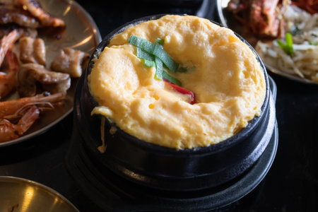 A fluffy steamed egg custard topped with green onion and chili, served in black stone bowl alongside seafood and side dishesの写真素材