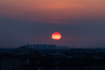 A red sun setting over urban skyline at dusk with layered clouds and warm sky