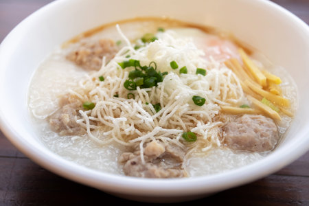 A rice porridge topped with crispy rice noodle, minced pork meatball, sliced ginger and chopped green onion in bowl, comforting savory breakfastの写真素材