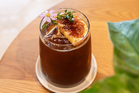 An iced coffee with caramelized orange slice and edible flower garnish on wooden table, refreshing...