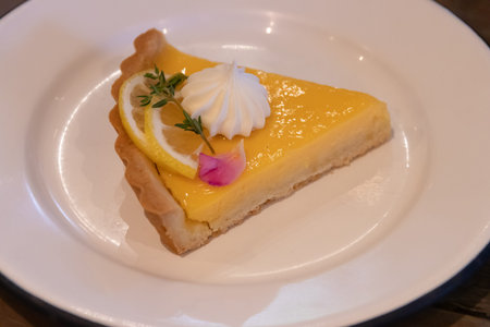 A lemon tart slice with whipped meringue, lemon slice and herb garnish on white plate, bright...