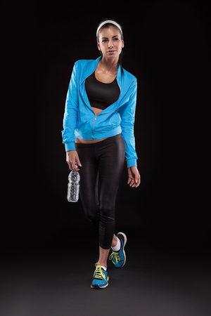 young brunette woman in jogging shoes after sportive exercise and holding the bottle of watter isolated on black  colored, retouched の写真素材