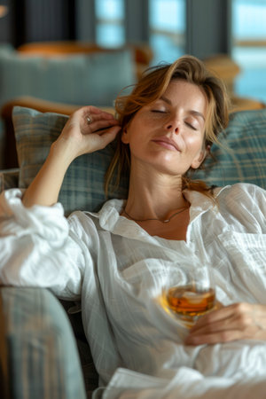 Serene Young Woman Relaxing on a Cozy Sofa with a Glass of Amber Beverage, Enjoying Peaceful Moments Indoor, Comfortable Home Setting AI Generativeの素材