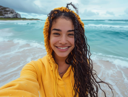 A joyful young woman in a vibrant yellow hoodie captures a moment of pure bliss as she takes a selfie against the backdrop of a picturesque beach Despite the overcast sky, her infectious smile reflects the vibrant beauty of the ocean&#39;s expanse, creating a captivating image of connection to nature and unbridled happiness AI Generativeの素材