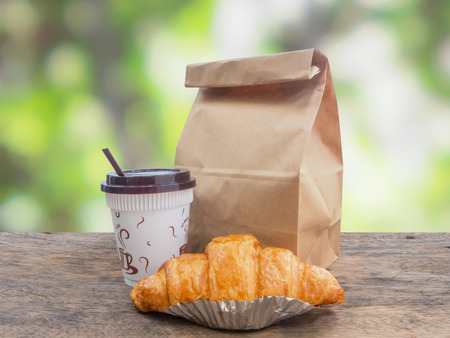 Breakfast to go,coffee and croissant with paper bag on wooden table over green defocused backgroundの写真素材