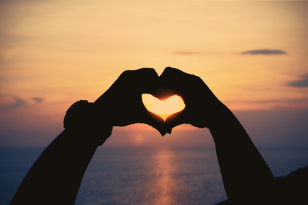 Silhouette human hands in heart symbol shaped love concept with sunlight and flare at sunset beach, vintage filter effectの写真素材